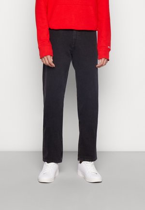 Henkilö, jolla on mustat suorat farkut, punainen pitkähiha-paita ja valkoiset tennarit, seisoo harmaalla lattialla valkoisen seinän edessä.