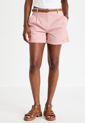 Frau, die hellrosa Shorts mit umgekrempeltem Saum, einem geflochtenen braunen Gürtel und braunen Riemchensandalen trägt, steht mit den Händen in den Taschen.