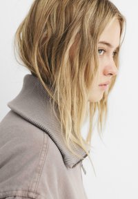 Krave, lys brun sweatshirt med ribbet tekstur og lynlås, båret af en person med langt, bølget blond hår.