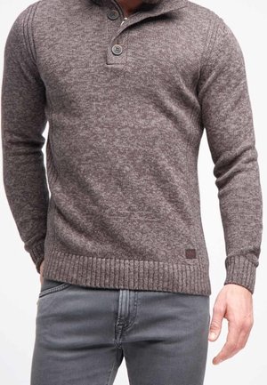 Pull en tricot marron avec un col montant, deux boutons et des poignets côtelés. Porté par-dessus un jean gris, avec un tissu texturé.