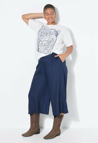 Witte t-shirt met zwart draaidesign, marineblauwe wijde broek en bruine cowboylaarzen. Gladde stof en comfortabele pasvorm met zijzakken.