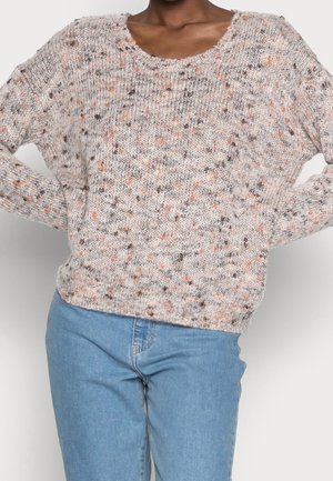 Loose-fit knit sweater with orange, brown, and cream speckles, worn with blue jeans, arms bent and hands on hips.