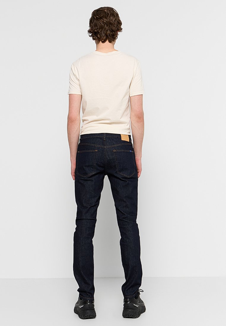 Dunkelblaue Jeans mit Slim Fit, ausgestattet mit einem braunen Lederpatch auf der Rückseite und kontrastierenden Nähten. Kombiniert mit einem hellbeigen T-Shirt.