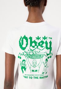 Hvit bomull t-skjorte med grønt grafisk design på ryggen, som viser stiliserte figurer, musikk-elementer, og teksten "Obey" og "Submit to the Rhythm."