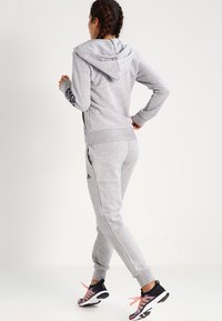 Survêtement de sport gris avec capuche, doté d'une fermeture éclair et de poches latérales, associé à des chaussures de course noires avec des lacets roses.