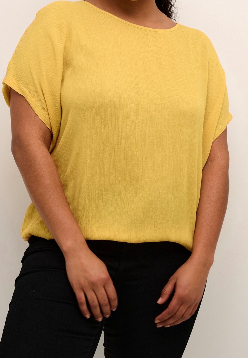 Femme portant un haut jaune ample à manches courtes et un pantalon noir, debout les mains détendues devant.