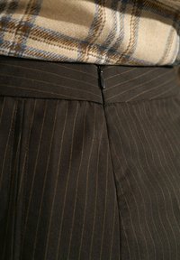 Gros plan d'une jupe marron à fines rayures avec une fermeture éclair noire visible et une partie d'un haut beige à carreaux rentrée à l'intérieur.