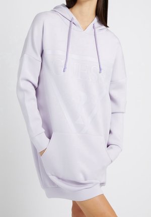 Kobieta ubrana w dużą, jasnofioletową sukienkę-bluzę z przednią kieszenią i subtelnym tłoczonym logo "Guess", trzyma ręce w kieszeniach.