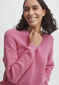 Rosa Strickpullover mit einem strukturierten Waffelmuster, langen Ärmeln und einem weiten Halsausschnitt. Gerippte Bündchen betonen das Design.