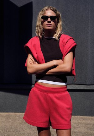 Mujer con pelo rubio rizado lleva gafas de sol negras, camiseta negra y pantalones cortos rojos con una sudadera roja sobre los hombros, de pie al aire libre.