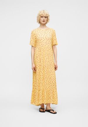 Femme aux cheveux blonds bouclés portant une robe jaune longue jusqu'aux chevilles avec des motifs floraux rouges et des sandales tongs noires, se tenant devant un fond blanc.