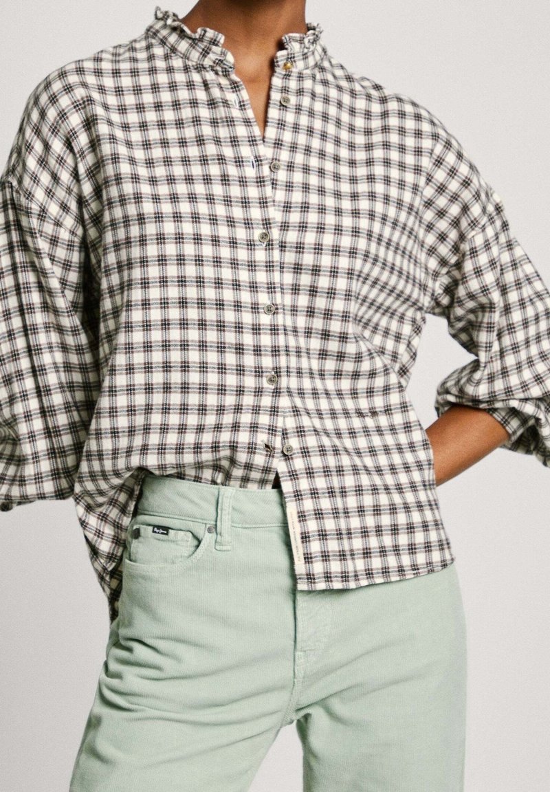 Personne portant une chemise ample blanche et noire à carreaux avec un col volanté et un pantalon taille haute vert clair, main sur la hanche.