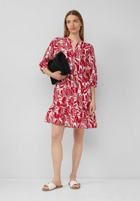 Rotes Kleid mit weißem abstraktem Blattmuster, dreiviertelÄrmel und gestuftem Saum. Kombiniert mit einer schwarzen, strukturierten Clutch und weißen Sandalen.