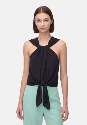 Donna che indossa un top nero senza maniche a halter annodato in vita con pantaloni verde chiaro a vita alta, in piedi su uno sfondo semplice.