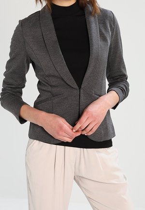 Person wearing a fitted gray blazer over a black turtleneck paired with loose, light-colored trousers, hands clasped in front.