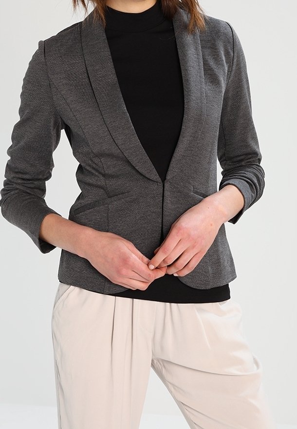 Personne portant un blazer gris ajusté par-dessus un col roulé noir, associé à un pantalon ample de couleur claire, les mains jointes devant.