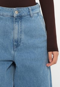 Jeans de denim azul claro con cintura alta, que cuentan con un cierre de botón y bolsillos laterales. La tela tiene una apariencia ligeramente texturizada y desgastada.