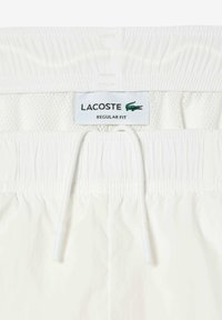 Balti šortai su tinklelio juosmeniu, elastinga raišteliu ir Lacoste logotipo etikete, ant kurios mėlyna spalva užrašyta "REGULAR FIT".