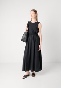 Svart langermet maxikjole med sammensunket liv og jevn tekstur, kombinert med svart lærveske og slip-on sandaler med blokkhel.
