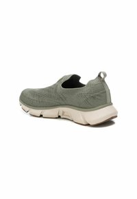 Zapatilla deportiva tipo slip-on en material de punto verde apagado con diseño texturizado y suela beige de contraste. Cuenta con una pequeña asa trasera.