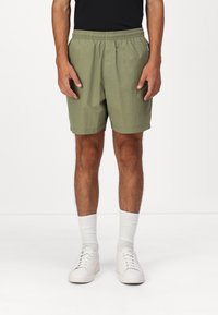 Shorts verdes oliva con cinta elástica en la cintura y bolsillos laterales, hechos de tejido ligero. Combinados con zapatillas blancas y calcetines.