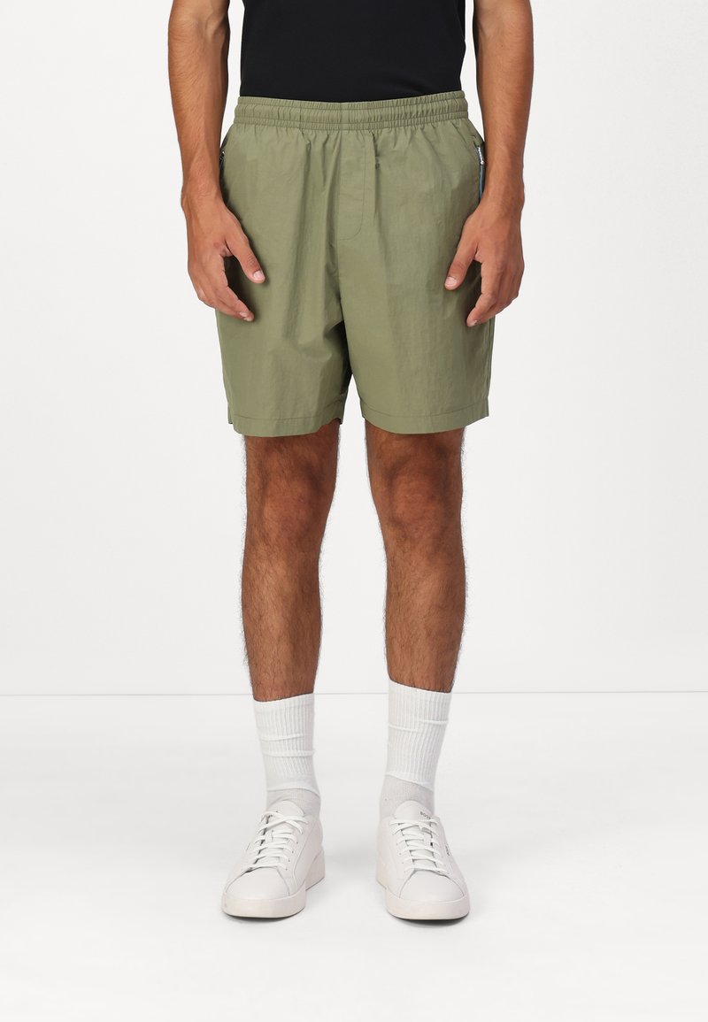 Shorts verdes oliva con cinta elástica en la cintura y bolsillos laterales, hechos de tejido ligero. Combinados con zapatillas blancas y calcetines.