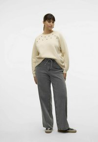 Bēšs sporta krekls ar smalkiem melniem akcentiem, kombinēts ar platām pelēkām džinsu biksēm. Audums izskatās mīksts; džinsi ir brīva, ikdienišķa stila.