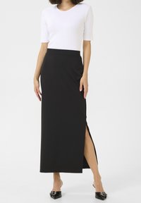 Jupe noire longueur cheville avec une fente latérale, en tissu lisse, associée à un haut blanc ajusté. Accentuée par des chaussures simples et élégantes.