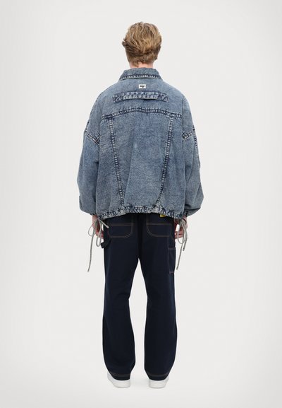 Jachetă oversized din denim de culoare bleu deschis, cu tiv desfăcut, talie ajustabilă cu șnur și detalii de cusătură în contrast. Purtată cu pantaloni negri.