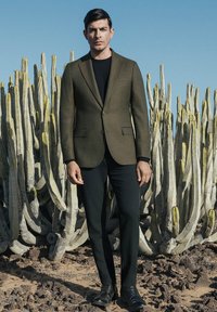 Zielona oliwkowa marynarka dopasowana z dwoma guzikami, w połączeniu z czarną koszulą i slim-fit czarnymi spodniami. Stoi wśród wysokich, kolczastych kaktusów.