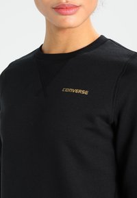 Primo piano di una persona che indossa una felpa nera con logo dorato "Converse" sul petto, con in evidenza il collo e parte del viso.