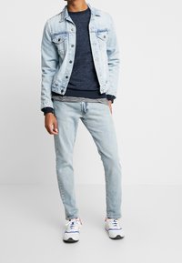 Chaqueta de mezclilla azul claro con dos bolsillos en el pecho, suéter de textura azul marino, jeans azul claro y zapatillas blancas con acentos rojos y azules.