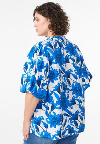 Florale Bluse mit einem lebhaften blauen Design auf weißem Hintergrund, kurzen puffärmel und einem lockeren Schnitt, aus leichtem Stoff gefertigt.