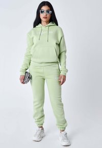 Sudadera con capucha de color verde claro, con bolsillo frontal y cordones, combinada con pantalones de chándal a juego. Ambos presentan sutiles acentos de logo. Zapatillas blancas.