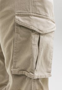 Vrecko na béžových nákladných nohaviciach s chlopňou, detailným prešívaním a skladaným dizajnom na bočnej strane nohavice na jednoduchom pozadí.