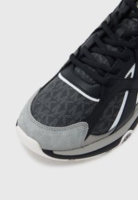 Černošedá teniska s texturovaným látkovým svrškem, semišovou špičkou a bílými detaily. Jsou vidět tkaničky a podpůrná podrážka.