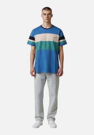 Jonge man die een blauw, groen, creme en zwart gestreept T-shirt draagt met lichtgrijze broek en witte sneakers, staand en gezicht naar voren gericht.