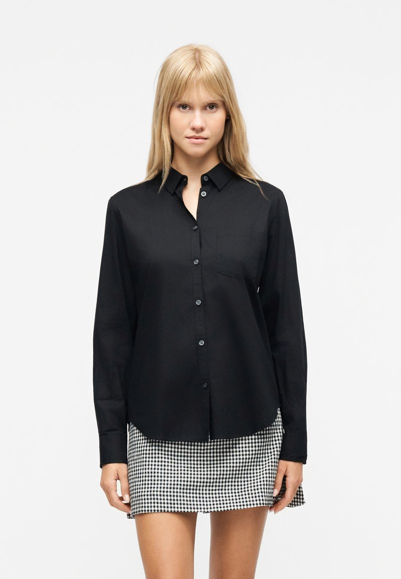 Γυναίκα φοράει μαύρο πουκάμισο με μακριά μανίκια και κουμπιά, με τσέπη στο στήθος, και μίνι φούστα με ασπρόμαυρο καρό σχέδιο, στέκεται μπροστά σε λευκό φόντο.