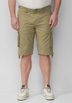 Man in beige cargoshorts en witte sneakers, staande tegen een lichtgrijze muur.