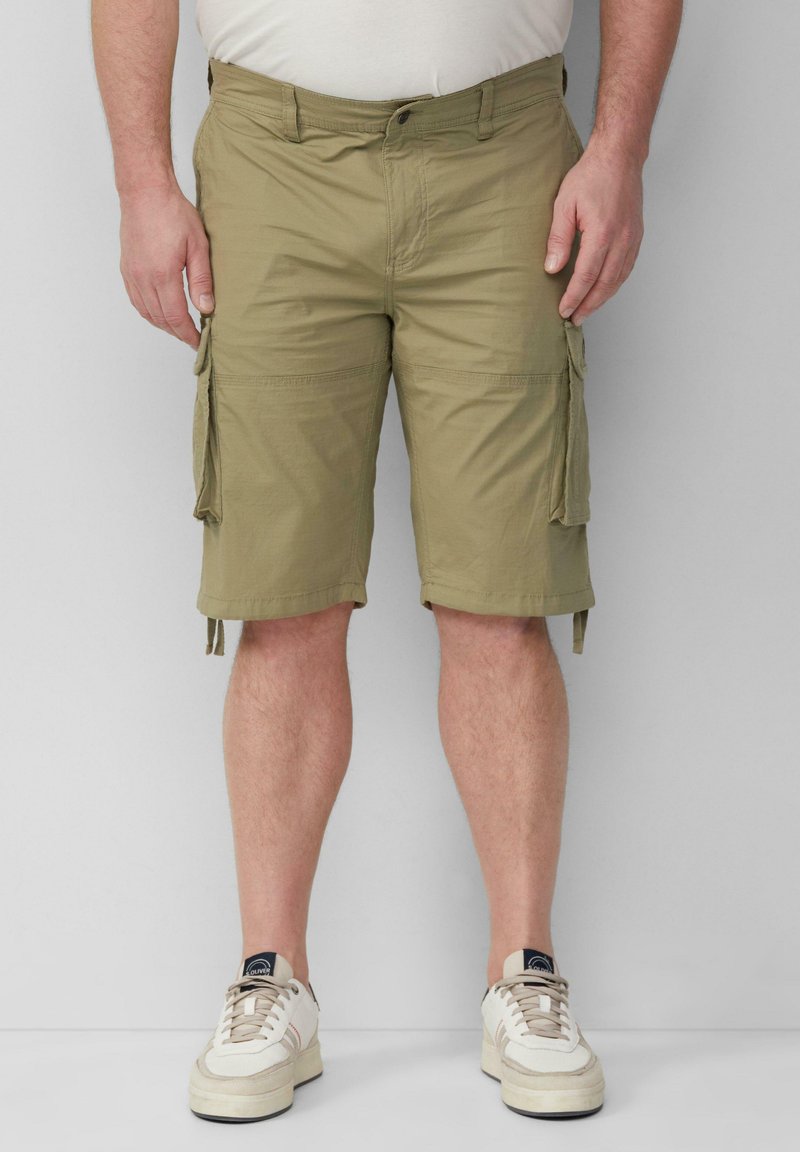 Man in beige cargoshorts en witte sneakers, staande tegen een lichtgrijze muur.