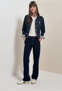 Tummansininen farkkutakki hopeisilla yksityiskohdilla, yhdistettynä tummiin naruhousuihin ja valkoisiin tennareihin. Malli seisoo vaaleataustalla.