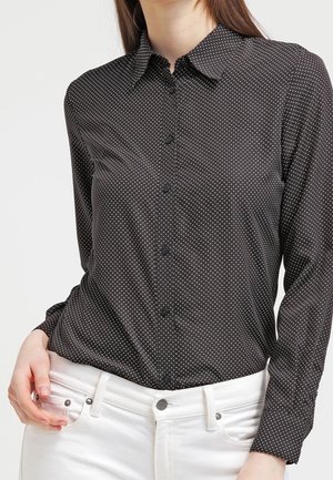 Femme portant une chemise noire à manches longues avec de petits pois blancs et un pantalon blanc, main posée sur la hanche.