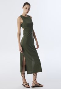 Olijfgroene mouwloze jurk met zijuitsparingen, halflang, met een slim fit en een zachte textuur. Gemaakt in combinatie met bruin sandaaltjes met bandjes.