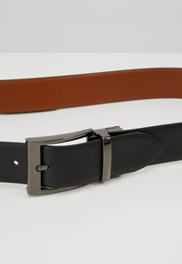 Cinturón de cuero negro y marrón con una correa texturizada negra, hebilla de metal plateado y un respaldo liso marrón, mostrando un diseño bicolor.