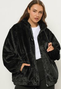 Czarna kurtka z sztucznego futra z zapięciem na zamek, kapturem i dużymi kieszeniami. Materiał ma puszystą teksturę i jest w luźnym kroju.