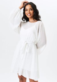 White dress made of sheer fabric with a tie waist. It features long, puffed sleeves and a textured pattern throughout.