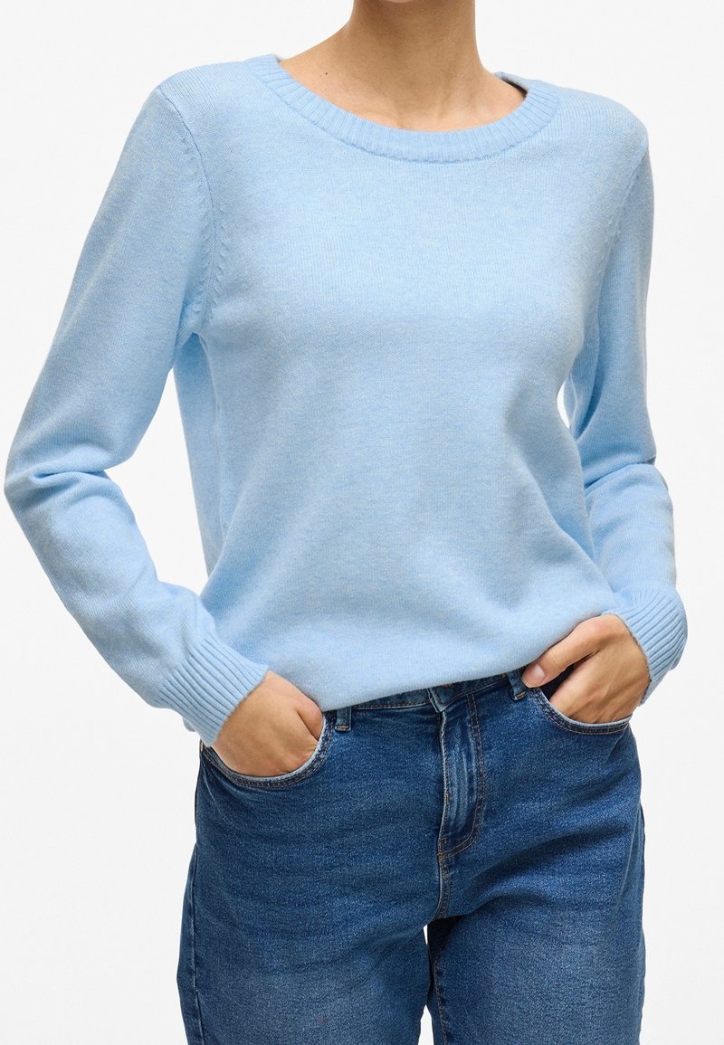 Personne portant un pull à manches longues bleu clair et un jean bleu, les mains dans les poches, sur un fond uni.