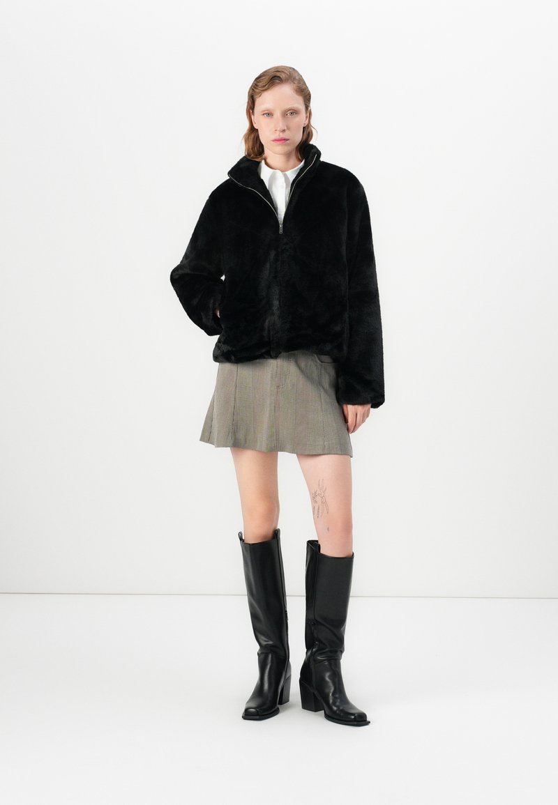 Veste en fausse fourrure noire avec fermeture éclair et col montant, associée à une jupe à carreaux grise et des bottes en cuir noir montantes. Texture lisse.