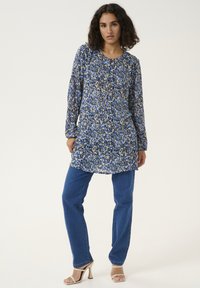 Tunique à manches longues en imprimé floral bleu et beige, avec un col rond et des détails de boutons, assortie à un jean droit bleu et des talons beiges.