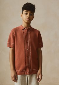 Polo a maniche corte color ruggine con lavorazione a maglia testurizzata, bottoni sul davanti e dettagli ricamati bianchi lungo la patta e le maniche.